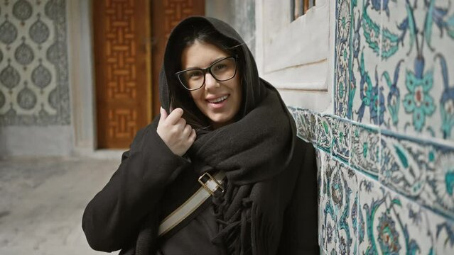 A smiling young woman with glasses, wearing a black shawl, stands against ornate blue tiles in istanbul's historical topkapi palace.