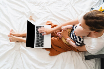 Mother working at laptop playing with baby in bed at home.