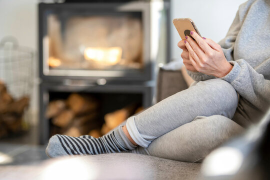 Young Woman sitting on the sofa in a clean modern home with his phone browsing with a warm fire in the background 