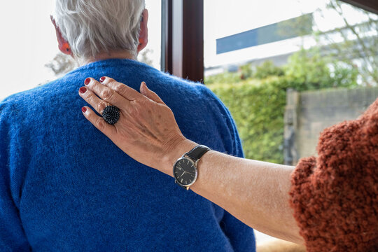 Two elderly friends standing looking out of the window consoling one another