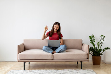 Fototapeta na wymiar Cheerful asian woman using laptop for video call, sitting cross-legged on sofa