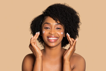 Happy black woman applying facial cream on cheeks and smiling