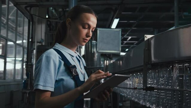 Medium side footage of young female inspector wearing polo shirt and jumpsuit standing near factory conveyor with tablet and controlling quality of PET bottles production at drinking water factory