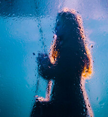View through a glass window with raindrops on a blurred silhouette of a girl on a autumn city street after rain against the bokeh of  city lights, night street scene. Focus on raindrops on glass	