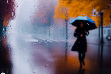 View through a glass window with raindrops on a broken silhouette of a girl with an umbrella walking along after rain ,  Focus on raindrops on glass	
