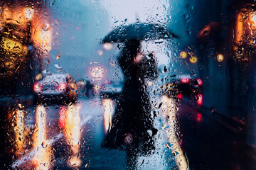 View through glass window with rain drops on blurred reflection silhouettesof a man andgirl in walking on a rain under umbrellas and bokeh city lights, night street scene. Focus on raindrops on glass	