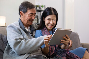 A senior couple in their 60s spends their free time sitting on a tablet, relaxing and watching entertainment programs, relaxing happily together on the sofa in the living room.