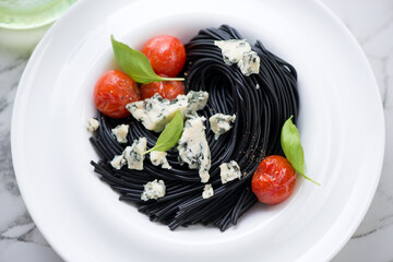 Squid-ink spaghetti served with blue cheese, roasted cherry tomatoes and green basil in a white plate, middle close-up, horizontal shot