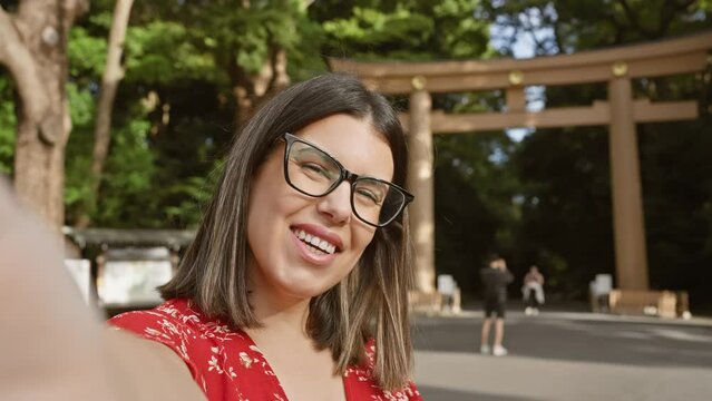 Gorgeous brunette hispanic woman in glasses waving to camera during exciting video call from meiji temple, tokyo