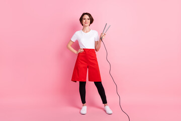 Full length body size photo of hairdresser keeping hot hair straightener smiling isolated on pastel pink background