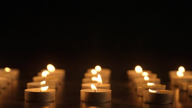 Rows of burning candles. The candles burn against a black background and the flames move when the wind blows. The concept of a memorial day. Background for advertising and design projects.
