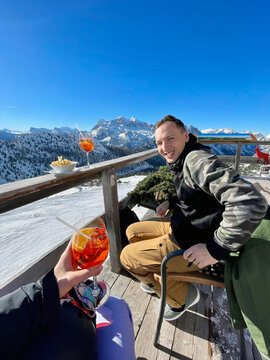 A handsome young man enjoying a break against the backdrop of mountains after snowboarding, sipping on an apres ski drink. Alpine scenery, mountain view.