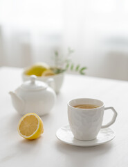 Fototapeta na wymiar A cup of tea with lemon and a teapot on a white table against the background of a kitchen window.