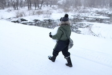 Fototapeta na wymiar A boy carries a ball of snow across a bridge over a river