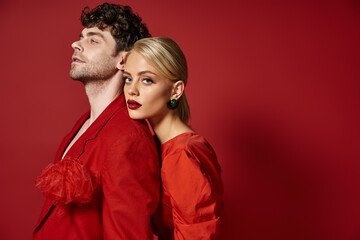 blonde woman with red lips leaning on shoulder of man in vibrant attire of red background, fashion