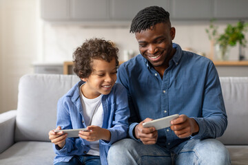 Joyful black father and son playing video games on cellphones together