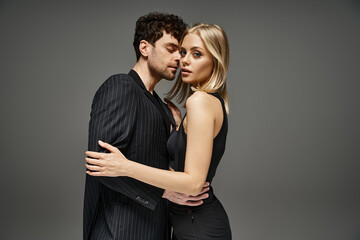 elegant woman in black dress seducing good looking man in evening attire on grey background, couple