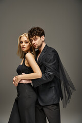 stylish couple in elegant attire, man in designer wear embracing woman in sleek black dress on grey