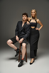 sophisticated couple in fashionable attire, woman in elegant black dress near man sitting on stool