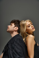 blonde woman sitting back to back with handsome man in pinstripe blazer on grey backdrop, couple