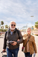 Vertical smiling Caucasian mature tourist couple standing posing looking at camera in street with their luggage. Husband and wife enjoying their retirees holidays wearing winter clothes on sunny day