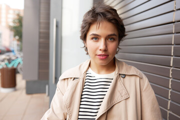 Outdoor portrait of smiling youthful stylish female city tourist in trench posing against textured background of building, walking in downtown on warm spring day, spending her weekend