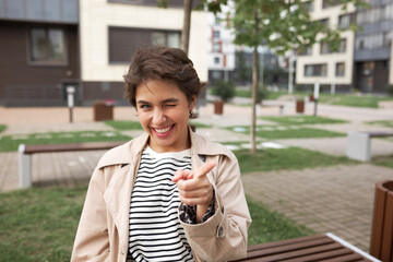 Outdoor portrait of winking woman pointing index finger at camera giving sign its you turn to...