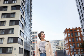 Outdoor portrait of young woman standing among mutli-storey buildings, admiring beauty and comfort of new residential area and being happy about making right choice to buy apartment in this place