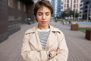 Pretty young woman standing with crossed hands and closed eyes, smiling relaxing walking in her neighborhood, dreaming of ideal life making wish of buying property in new residential area