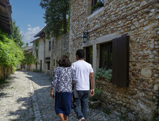 Fototapeta na wymiar Couple qui se promène en été dans une ruelle pavée d'un village médiéval, la rue est bordée de murs en pierres avec des fleurs aux fenêtres 
