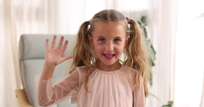 Positive preteen girl with long blond ponytails looking at camera while standing in blurred living room and waving hand in daylight against curtain and green plant