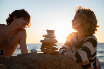 young cute guy looks lovingly at his girlfriend while sitting on beach at sunset. in front of them stands a balancing tower of stones. concept of love, relaxation, rest. Spending time together
