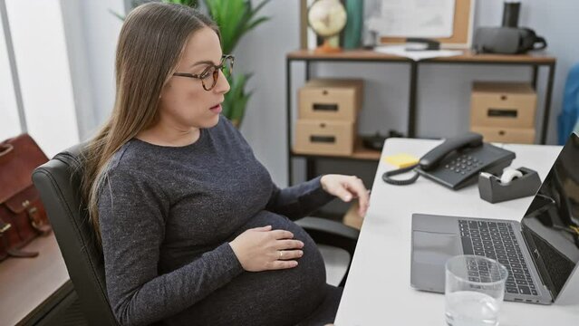 A pregnant hispanic woman experiencing discomfort at her office desk, surrounded by work equipment.