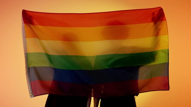 Silhouettes of people with large flag on yellow background. Silhouettes of a man and a woman with LGBT pride flag holding in hands.