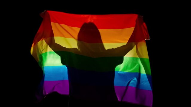 Person silhouette holding rainbow flag against black background. Silhouette of woman with LGBT pride flag holding in hands.