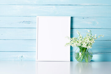 Lily of the valley flowers in glass vase, blank photo frame on wooden blue background.