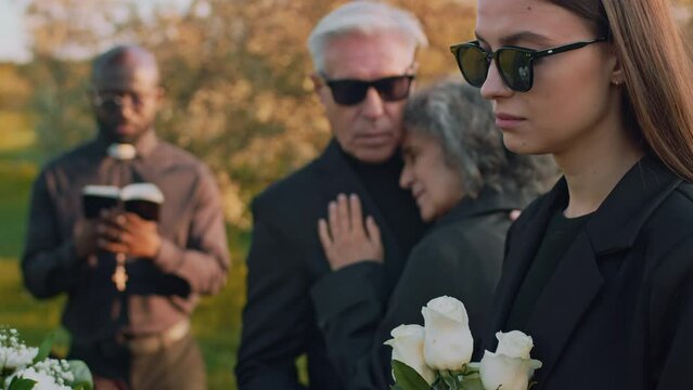 Side tilt closeup of Caucasian young lady looking painfully at wooden casket with white roses in hands at outdoor funeral ceremony