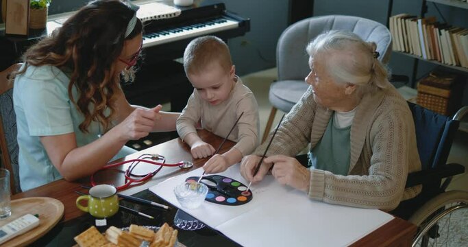 A little boy and his elderly grandmother with disability, who is cared for by a nurse or caregiver, spend their free time painting together at home