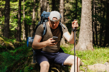 Adult man with smartphone backpack trekking stick sitting in green forest