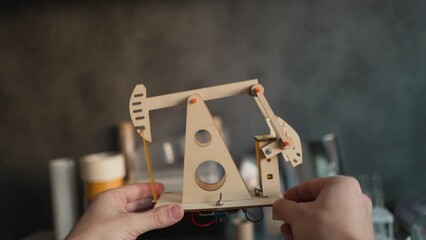 POV video of hands raising a working wooden oil pumpjack model with motor at eye level on a desk for inspection