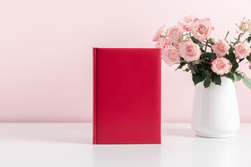 Red book mockup, diary, cover, white table, pink roses on pink background. Front view. Place for text, copy space, mockup
