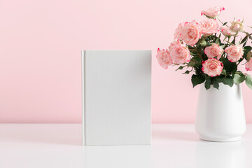 White book mockup, diary, cover, white table, pink roses on pink background. Front view. Place for text, copy space, mockup