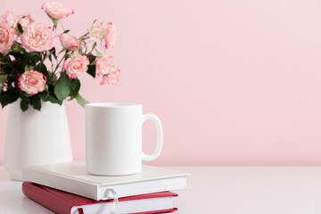White cup mockup, stack of notebooks and bouquet pink roses in vase on pink table background. Front view. Place for text, copy space, mockup