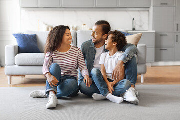 Smiling black family of three sitting on floor in living room - obrazy, fototapety, plakaty