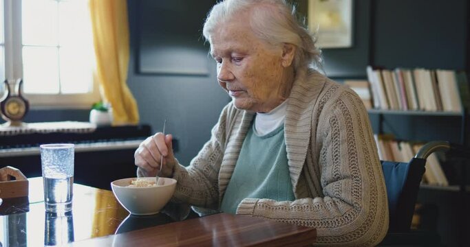 Lonely old woman in wheelchair sitting and eating noodles during lunch at home