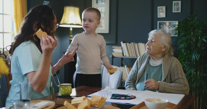 Happy elderly grandmother with disability in a wheelchair with grandson and home caregiver smiling painting and eating together in living room at home