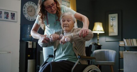 Female nurse physiotherapy worker help elderly woman with disability to exercise with dumbells, caregiver assist a senior woman in wheelchair to do physical therapy and exercise at home