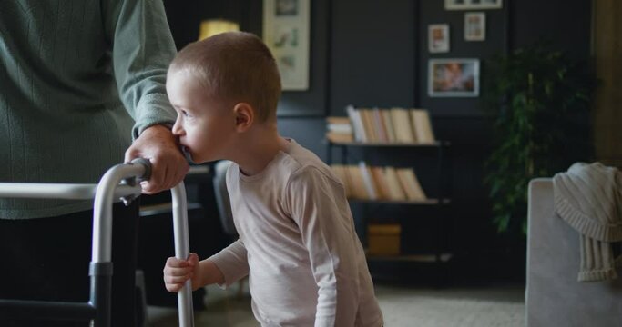 Little boy grandson kisses old grandmother's hand giving her help and support to walk using a walker at home