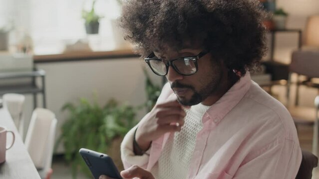 Waist up of young Biracial freelance man with long curly messy hair scrolling on smartphone while drinking coffee, sitting at desk in creative home office