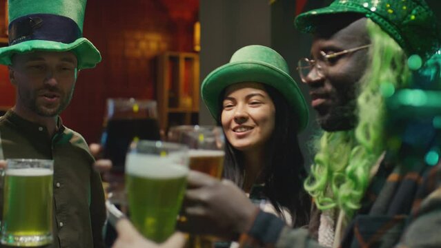 Chest up panning shot of multiracial adult friends wearing green hats and clanking glasses of beer while on party celebrating St Patricks Day together in bar on March 17